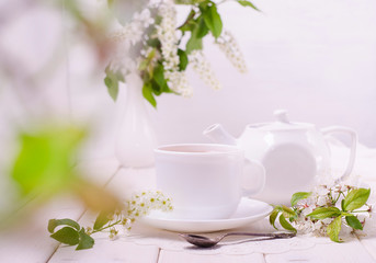 Obraz na płótnie Canvas Tea ceremony. White teapot and cup on a white wooden table with flowers and with a bokeh of green leaves. Herbal tea.