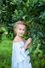 Little girl with a pigtail on her head is worth in apple orchard. Gardening. Childhood concept. Child walks in woods in spring day. Summer holidays. Portrait young girl outdoors. Child relax in park