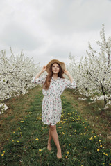 Obraz premium Stylish girl walks in the cherry orchard. Young woman in a hat and dress on the nature between flowering trees.
