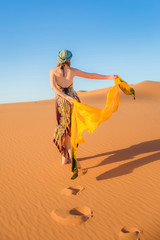 Beautiful young slim woman in a multi-colored dress with a yellow scarf, in a turban and sunglasses poses at dawn in the Sahara desert. Morocco Fashion.