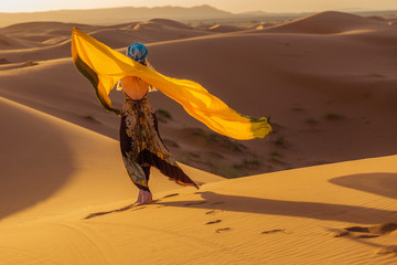 Belle jeune femme mince dans une robe multicolore avec une écharpe jaune, dans un turban et des lunettes de soleil pose à l& 39 aube dans le désert du Sahara. Mode marocaine.