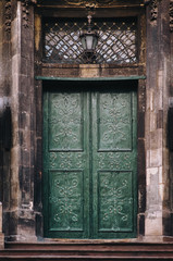 The old vintage green wooden door on the facade of the baroque Dominican Cathedral in Lviv is decorated with decorative forged flowers. Lamp and lattice window above the doorway.