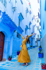 A beautiful caucasian woman in a yellow dress is dancing in a street in a blue city in Morocco. Chefchaouen.