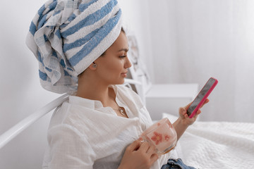 Cute pretty girl is wearing towel on her head chilling at home with phone and cup of tea. Beauty and cosmetics concept.