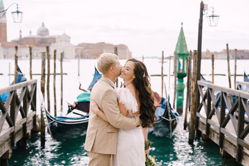 Fototapeta na wymiar The bride and groom kiss on the gondola pier, hugging, in Venice, near St. Mark's Square, overlooking San Giorgio Maggiore and the sunset sky. The largest gondola pier in Venice, Italy.