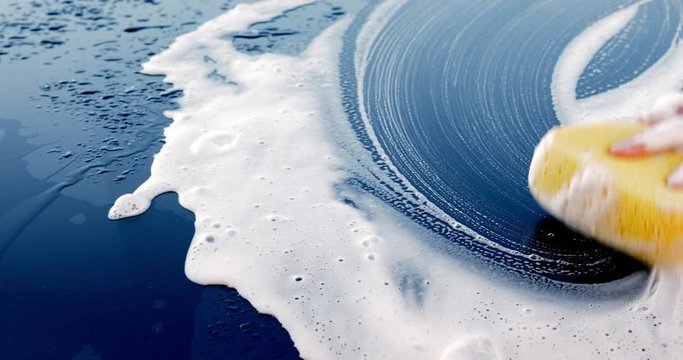 Washing car with sponge and shampoo, hand spreading foam on wet dark car close-up, 4k