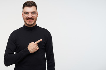 Glad young attractive dark haired bearded male smiling widely at camera while showing aside with index finger, standing over white background in black sweater