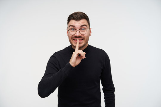 Portrait of excited young brown haired bearded man keeping raised forefinger on his mouth and raising surprisedly eyebrows while standing over white background