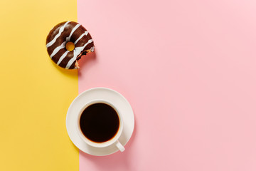 Flat lay composition with a bitten chocolate donut and a cup of coffee on a yellow-pink background with a copy space