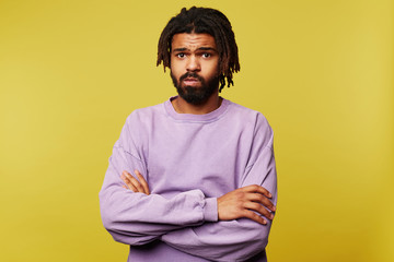 Indoor photo of young attractive dark skinned brunette guy with dreadlocks keeping his hands folded while looking sadly at camera, posing over yellow background