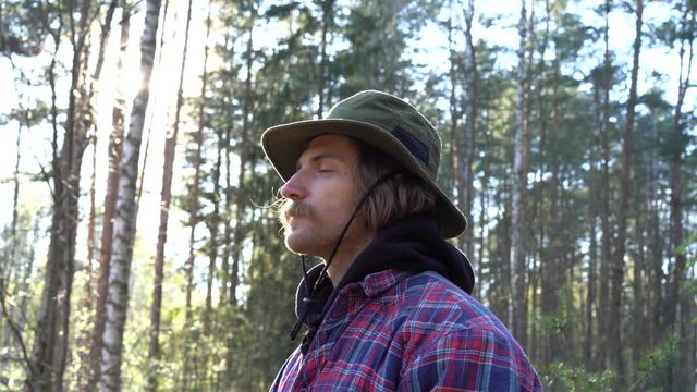a nice young european hipster guy with mustache in a checkered shirt and a green hat enjoys freedom, breathes fresh air in quiet forest 