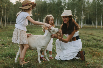 Fototapeta premium Mother and daughters feed a goat in a meadow in white dresses on a farm summer day