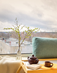 Spring twigs in the bottle on the table with a plaid, pillow, book, teapot and a cup at the window with a rainy city outside the window (on the background). Concept of a feeling of coziness at home.