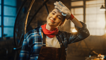 Obraz na płótnie Canvas Young Beautiful Empowering Woman Rubs Her Forehead and Gently Smiles at the Camera. Authentic Fabricator Wearing Work Clothes in a Metal Workshop.