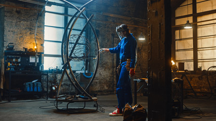 Young Contemporary Female Artist in Blue Jumpsuit and Safety Mask is Fitting a Metal Ring to a Tube Sculpture in a Studio Workshop. Empowering Woman Makes Modern Brutal Abstract Artwork Out Of Steel.