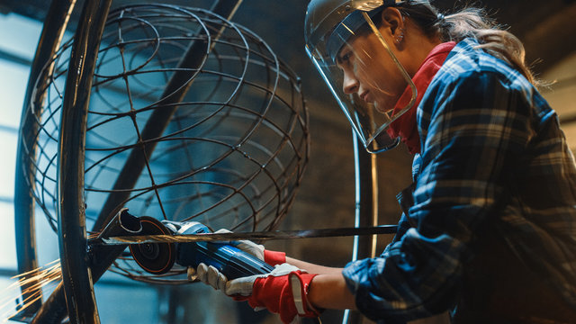 Close Up of Young Female Fabricator in Safety Mask. She is Grinding a Metal Tube Sculpture with an Angle Grinder in a Studio Workshop. Empowering Woman Makes Modern Abstract Metal Artwork.