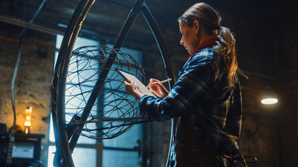 Beautiful Female Artist Sketches on a Tablet Computer Next to Brutal Metal Sculpture in Studio. Tomboy Girl Wears Checkered Shirt and Apron. Contemporary Fabricator Creating Abstract Steel Art.