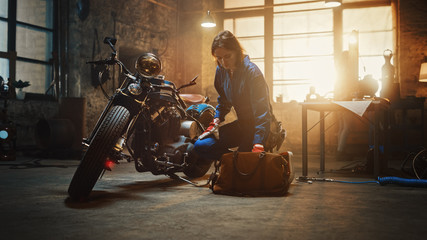 Young Beautiful Female Mechanic Comes Working on a Custom Motorcycle in Garage. Talented Girl Wearing a Blue Jumpsuit. She Looking for an Instrument in a Bag. Creative Authentic Workshop.