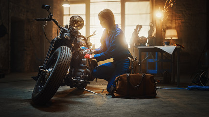 Young Beautiful Female Mechanic Comes Working on a Custom Motorcycle in Garage. Talented Girl Wearing a Blue Jumpsuit. She Uses a Ratchet to Tighten Nut Bolts. Creative Authentic Workshop.