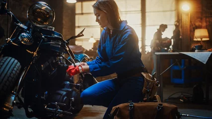 Papier Peint photo autocollant Moto Young Beautiful Female Mechanic is Working on a Custom Bobber Motorcycle. Talented Girl Wearing a Blue Jumpsuit. She Uses a Spanner to Tighten Nut Bolts. Creative Authentic Workshop Garage.