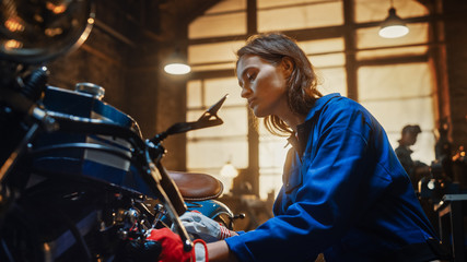 Fototapeta na wymiar Young Beautiful Female Mechanic is Working on a Custom Bobber Motorcycle. Talented Girl Wearing a Blue Jumpsuit. She Uses a Spanner to Tighten Nut Bolts. Creative Authentic Workshop Garage.