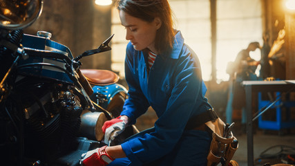 Young Beautiful Female Mechanic is Working on a Custom Bobber Motorcycle. Talented Girl Wearing a Blue Jumpsuit. She Uses a Ratchet Spanner to Tighten Nut Bolts. Creative Authentic Workshop Garage.