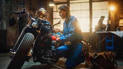 Fototapeta na wymiar Young Beautiful Female Mechanic is Working on a Custom Bobber Motorcycle. Talented Girl Wearing a Blue Jumpsuit. She Uses a Ratchet Spanner to Tighten Nut Bolts. Creative Authentic Workshop Garage.