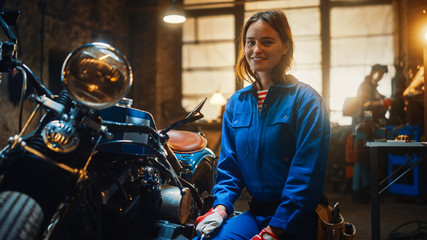 Fototapeta na wymiar Young Beautiful Female Mechanic is Working on a Custom Bobber Motorcycle. Talented Girl Wearing a Blue Jumpsuit. She Smiles at the Camera. Creative Authentic Workshop Garage.