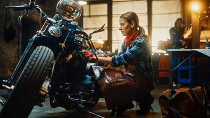 Obraz na płótnie Canvas Young Beautiful Female Mechanic is Working on a Custom Bobber Motorcycle. Talented Girl Wearing a Checkered Shirt and an Apron. She Uses a Spanner to Tighten Nut Bolts. Authentic Workshop Garage.