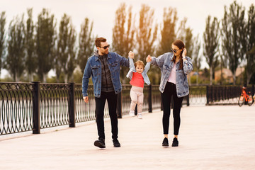 Young fashionable family walks along the embankment of the river, mom and dad hold daughter hands