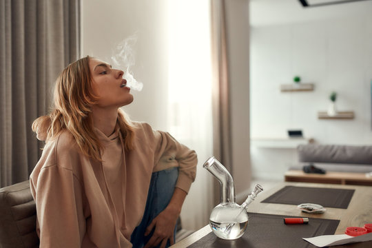Relax, renew, revive. Young caucasian woman exhaling the smoke while smoking marijuana from a bong or glass water pipe, sitting in the kitchen. Red weed grinder and lighter on the table
