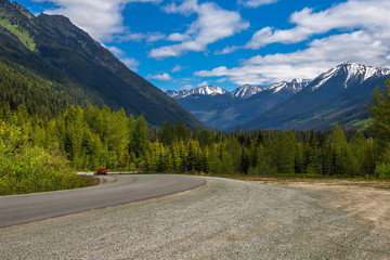 Fototapeta premium Highway from sea to sky. A car drives down a mountain road to the Lillooet Town, deciduous forest on the sides of the road a mountain range on the horizon against a blue cloudy sky