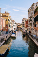 Fototapeta na wymiar Classic view of the narrow sea Venetian canal. Colorful buildings stand opposite each other, boats moored near shore, gondolier rides tourists on a gondola in Venice, Italy. Blue skies on a sunny day.