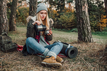 Hiking and travel along in the forest. Concept of trekking, adventure and seasonal vacation. Young woman with thermos sitting in woods.