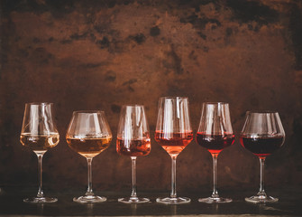 Various shades of Rose wine in stemmed glasses placed in line from light to dark colour on concrete table, rusty brown background behind, copy space. Wine bar, wine shop, wine tasting concept