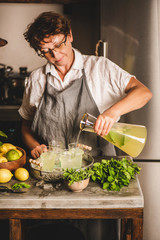 Elderly smiling woman making fresh homemade lemonade over kitchen counter. Woman pouring cold lemonade with mint and lemon into glasses with ice in rustic kitchen interior