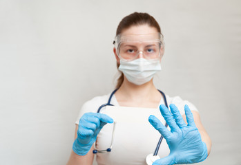 Female doctor in goggles shows the stop coronavirus sign. A young doctor in medical uniform with a protective face mask and gloved hand showing a stop sign