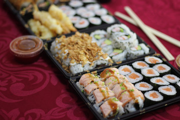 Sushi take-away delivery boxes with salmon Hosomaki, Californias, URAMAKI, fried onion, crispy onion, braised salmon, bowl of bittersweet, sweet and sour sauce, chopsticks on a red tablecloth