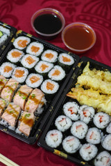 Sushi take-away delivery boxes with salmon Hosomaki, Californias, URAMAKI, breaded shrimp, tempura shrimp, bowl of bittersweet, sweet and sour sauce, chopsticks on a red tablecloth vertical
