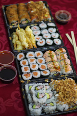 4 Sushi take-away delivery boxes with salmon Hosomaki, Californias, URAMAKI, fried onion, crispy onion, breaded shrimp, tempura shrimp, bowl of bittersweet, sweet and sour sauce, chopsticks on a red t