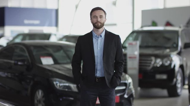 A young man in the auto dealership is posing. He is standing between new cars, looking at the camera and smiling.