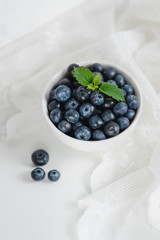 Fresh juicy blueberries with a sprig of mint in a white bowl with lace fabric on a white background.