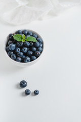 Fresh juicy blueberries with a sprig of mint in a white bowl with lace fabric on a white background.