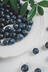 Fresh juicy blueberries in a white plate with green leaves on a gray background. 