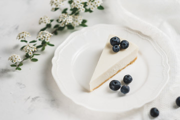 Blueberry cheesecake on a white plate, lace fabric and a branch with white flowers. 