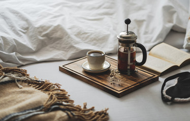 Coffee brewed in a French press and a cup on a wooden board with open book in bed. Morning at home, coziness and comfort. - Powered by Adobe