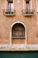 Fototapeta na wymiar Close-ups of building facades in Venice, Italy. A stone arch above a wooden window on the facade of building. White curtains in window, small flowers in pots on windowsill with a metal forged fence