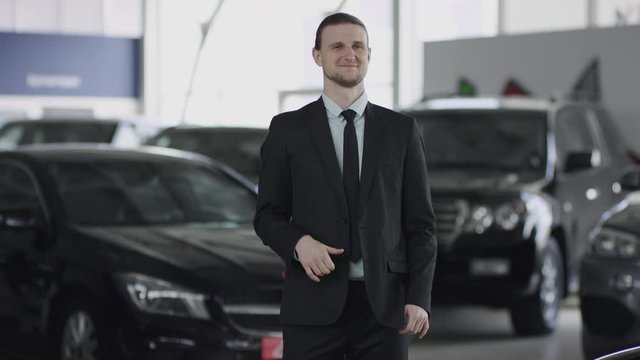 Standing between new cars in auto dealership man is giving thumb-up and posing in car showroom. He shows thumb-up looking at camera and smiling.
