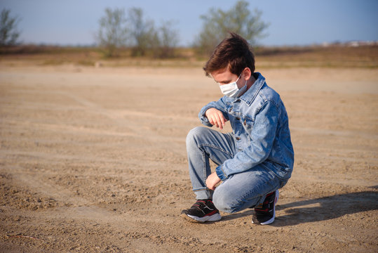 A boy of 8-9 years old in a protective mask and jeanse clothes outdoors. Kid is sitting on thr ground, upset and depressed due to the epidemic of coronavirus. Sunny weather. Pandemic covid 2020
