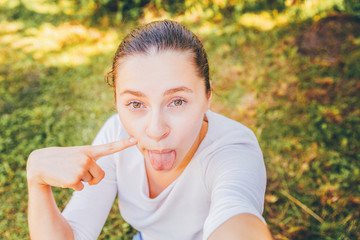 Fototapeta premium Young girl take selfie from hands with phone showing tongue and funny face sitting on park or garden background. Portrait of young attractive woman making selfie photo on smartphone in summer day.
